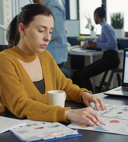 Portrait of startup employee comparing two sheets of paper with charts analyzing financial data while drinking morning coffee. Woman working in busy marketing department sitting at desk with laptop.